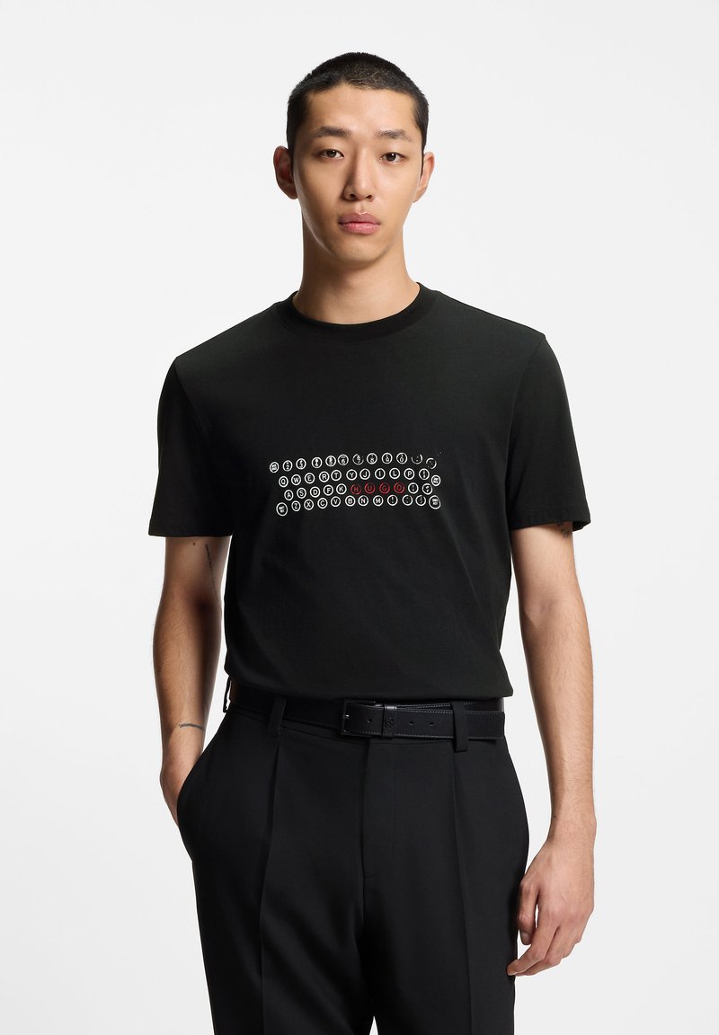 Crna pamučna majica s prednjim grafičkim prikazom bijelih i crvenih kružnih uzoraka. Kratki rukavi, okrugli izrez i opušteni kroj.