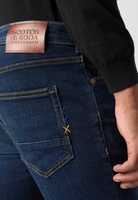 Pantalones vaqueros de denim azul oscuro con costuras naranjas, un parche de cuero en la parte trasera con "SCOTCH & SODA AMSTERDAM" y bordados decorativos en varios colores.
