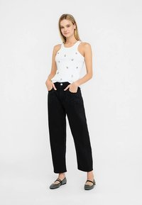 Canotta bianca con piccoli dettagli decorativi; pantaloni neri a vita alta con tasche frontali impreziosite da borchie; scarpe basse nere.