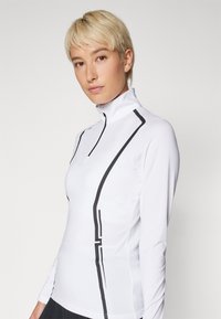 Λευκή αθλητική μπλούζα με μακριά μανίκια και ψηλό γιακά, χαρακτηρισμένη από μαύρες διαγώνιες ρίγες και φερμουάρ στον λαιμό. Κατασκευασμένη από λείο, ελαστικό ύφασμα.