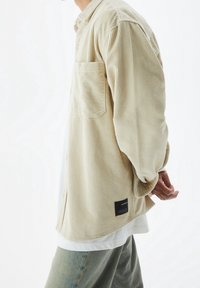 Chemise en velours côtelé beige avec col, manches longues et poche poitrine unique. Elle arbore une étiquette noire à l'ourlet, portée par-dessus une chemise blanche.