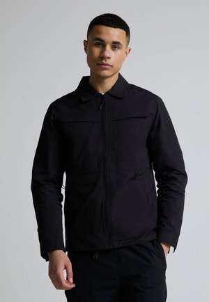 Jonge man met kort haar, gekleed in een zwart jack met rits en kraag, staand tegen een neutrale grijze achtergrond.