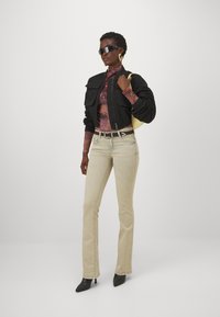 Veste bomber noire coupée, haut à manches longues violet, jean beige clair, escarpins noirs à talons pointus et sac jaune. Les accessoires comprennent des lunettes de soleil et des boucles d'oreilles.