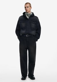 Svart denimjacka med bröstfickor, kombinerad med lösa svarta byxor. En grå bandana bärs under jackans huva. Klumpiga svarta skor.