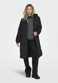 Manteau d'hiver noir long avec capuche, présentant des coutures verticales et des poignets élastiques. Porté sur un pull rayé et un pantalon noir.