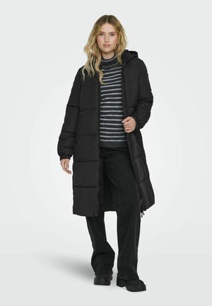 Manteau d'hiver noir long avec capuche, présentant des coutures verticales et des poignets élastiques. Porté sur un pull rayé et un pantalon noir.