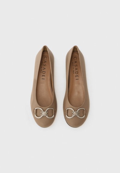 Chaussures plates beiges avec une finition en cuir lisse et un accent métallique argenté comportant des anneaux entrelacés au niveau des orteils. Design simple et classique.