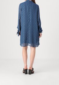 Femme portant une robe bleue transparente à manches longues avec des pois argentés, debout pieds nus sur un sol blanc devant un fond blanc.