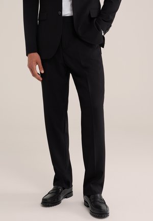 Bărbat purtând pantaloni negri de costum croiți pe măsură, pantofi negri eleganți și sacou negru, cu o mână în buzunar, pe un fundal neutru.