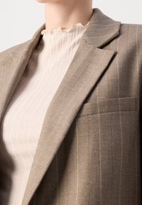Hnědý blazers s proužky z texturované látky. Má kapsu na levé straně a je vrstvený přes světle krémový top s jemným žebrováním.