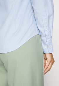 Hele sinine nööbiga särk pika varrukaga, valgete nööpidega ja kõverate äärtega, kombineeritud laia rohelise pükste paariga.