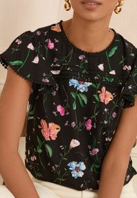 Blusa a maniche corte floreale nera, con fiori multicolori, bordi arricciati e una rifinitura in pompon neri attorno al collo e alle maniche.