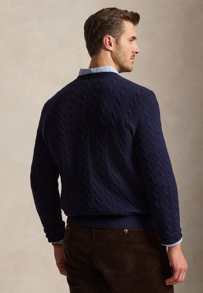 Uomo che indossa un maglione a trecce blu navy sopra una camicia azzurro chiaro con colletto, in piedi leggermente girato di lato su uno sfondo semplice.