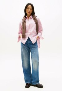 Lys pink og hvid stribet skjorte med knaplukning, afslappet pasform; parret med brede blå jeans og et brunt strikket tørklæde hængende om halsen.