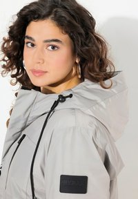 Giacca a vento grigia chiara con cappuccio, realizzata in materiale impermeabile e liscio. Presenta un cappuccio regolabile, dettagli neri e un'etichetta in tessuto sulla manica.