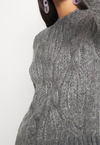 Šedý pletený svetr s texturovaným kabelkovým vzorem, kulatým výstřihem a ribovaným lemem. Tkanina vypadá měkce a silně.