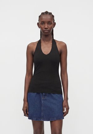 Femme aux cheveux tressés portant un haut noir sans manches à col halter et une jupe en jean bleue, debout devant un fond clair uni.