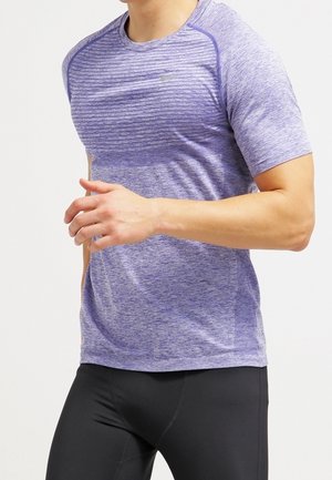 Homme portant un t-shirt de sport violet à manches courtes et un legging noir d'entraînement, montré du cou aux hanches, avec un bras plié.