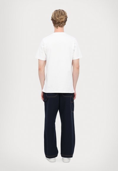 Baltas kokvilnas T-krekls ar īsām piedurknēm, skatīts no mugurpuses, savienots ar tumši ziliem platajiem biksēm ar redzamām vīlēm un kabatām.