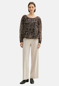 Schwarze durchsichtige Bluse mit floralem Muster, langen transparenten Ärmeln und gerafftem Halsausschnitt, kombiniert mit hellbeigen weit geschnittenen Hosen und schwarzen Loafers.