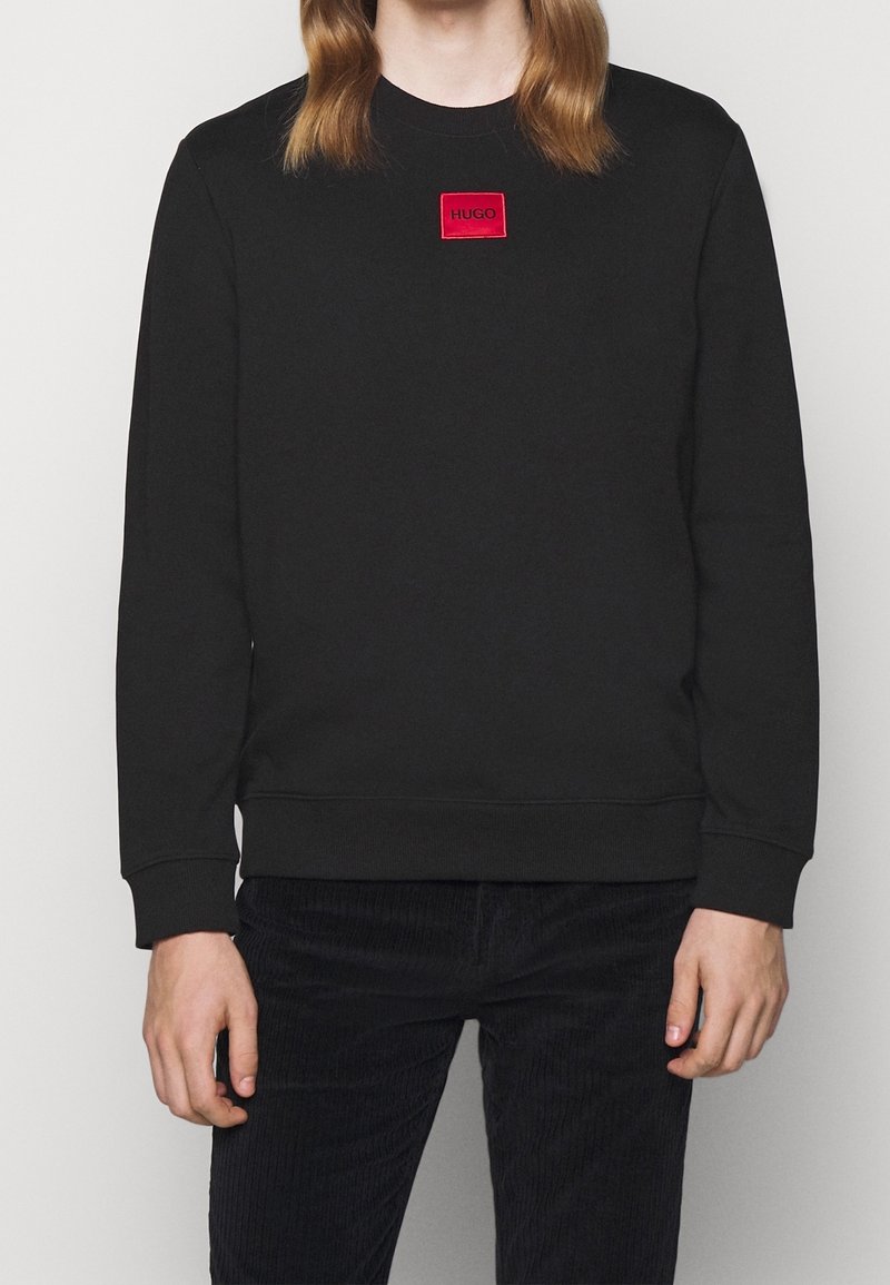 Sudadera negra con cuello redondo, que presenta un parche de logo cuadrado rojo en el pecho. Material de algodón con puños y dobladillo de canalé.