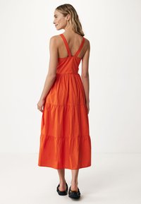 Oranžové vrstvené maxi šaty s nastavitelnými ramínky, přiléhavým korzetem a splývavým spodním dílem. Nosené s černými sandály na masivní podešvi.