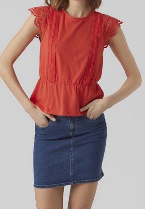 Mujer con una blusa roja sin mangas con mangas de encaje y una falda de mezclilla azul, con las manos en los bolsillos de la falda, sobre un fondo gris liso.