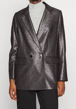 Persoon draagt een glanzend donker metallic colbert over een lichtgrijze coltrui en zwarte broek.