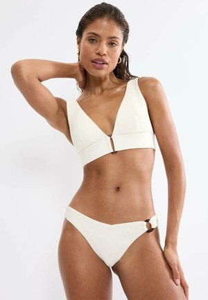 Woman posing in a white two-piece bikini with a ring detail on the bottom against a plain white background.