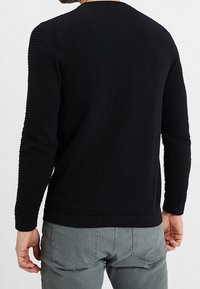 Černý pletený svetr s texturovanými žebrovanými detaily na ramenou a rukávech, kulatým výstřihem a přiléhavým spodním okrajem. Nosený s šedými kalhotami.