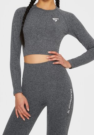 Žena v odpovídajícím šedém texturovaném dlouhém topu s krátkým rukávem a legínách s vysokým pasem, na stehně s nápisem "SQUATPROOF".