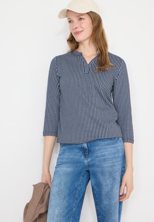 Kvinde iført en cremefarvet kasket, marineblå stribet langærmet skjorte, blå jeans, holder en beige jakke mod en hvid baggrund.