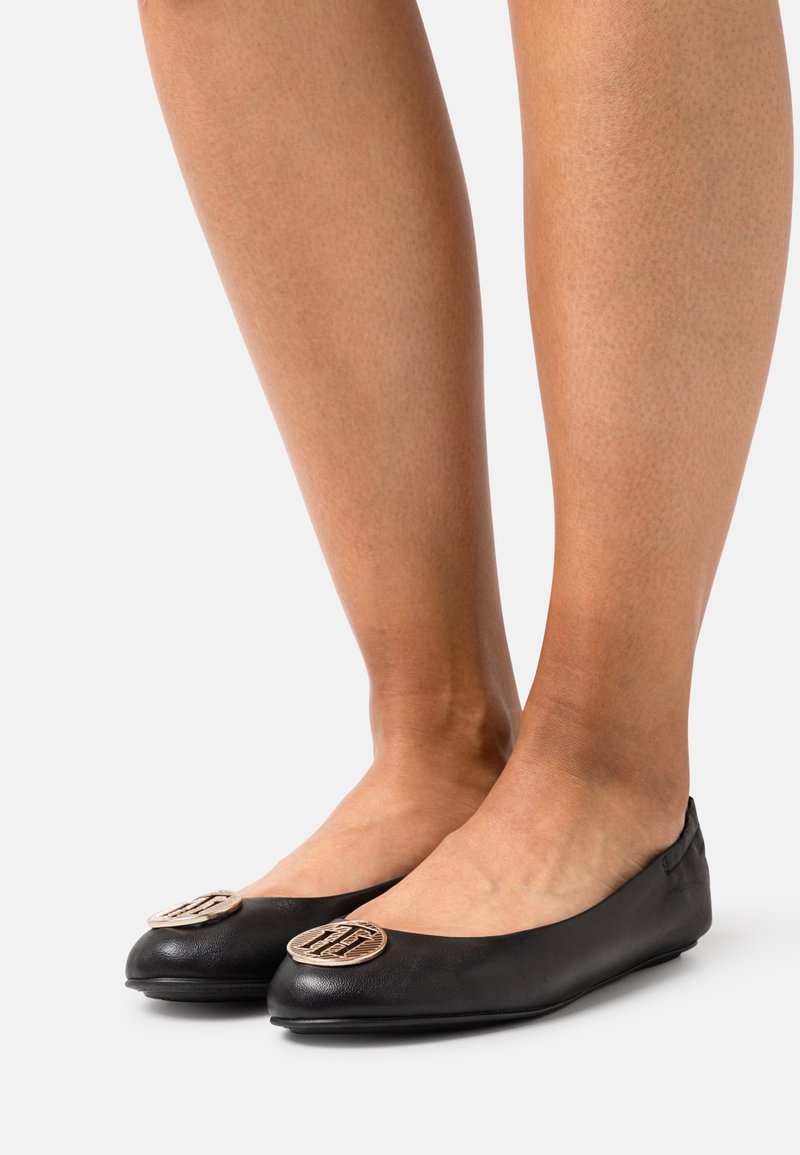 Pieds portant des ballerines en cuir noir avec des médaillons décoratifs ronds dorés sur les orteils sur un fond clair uni.