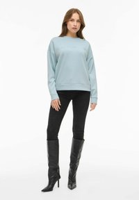 Moteris vilkinti šviesiai mėlyną džemperį su užrašu "MINIMALIZMAS", juodas slim fit džinsus ir juodas aukštas kulnų batus, stovinti prieš baltą foną.