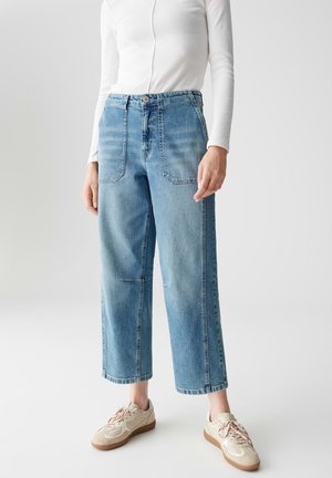 Personne portant un jean large court taille haute bleu clair, un top blanc à manches longues et des baskets beiges à lacets sur un fond uni.