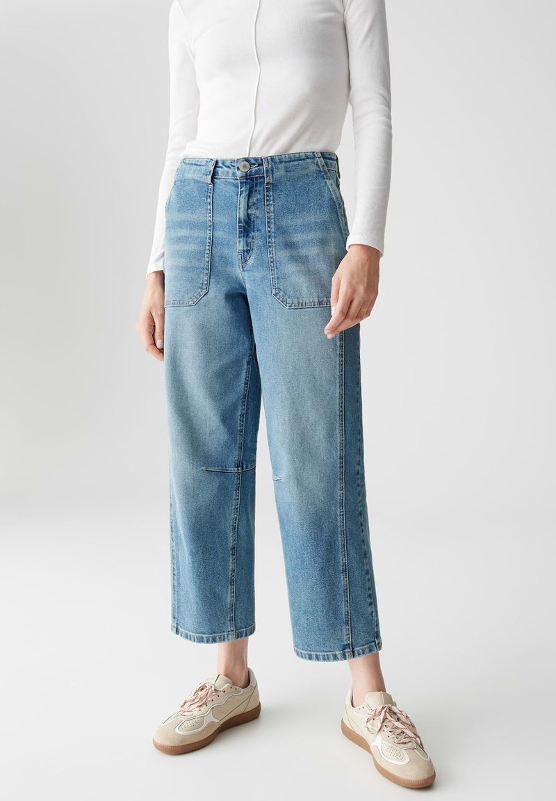 Personne portant un jean large court taille haute bleu clair, un top blanc à manches longues et des baskets beiges à lacets sur un fond uni.