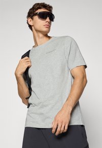 Junger Mann, der übergroße schwarze Sonnenbrille, ein hellgraues T-Shirt und schwarze Shorts trägt, während er einen schwarzen Rucksackriemen über einer Schulter hält.