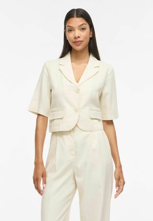 Femme aux longs cheveux foncés portant un blazer crème à manches courtes boutonné et un pantalon taille haute assorti, debout devant un fond blanc.