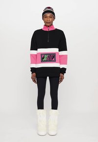 Hanorac colorat în negru și roz, cu guler înalt, închidere cu fermoar și patch cu logo. Asortat cu colanți negri și cizme căptușite cu blană albă.