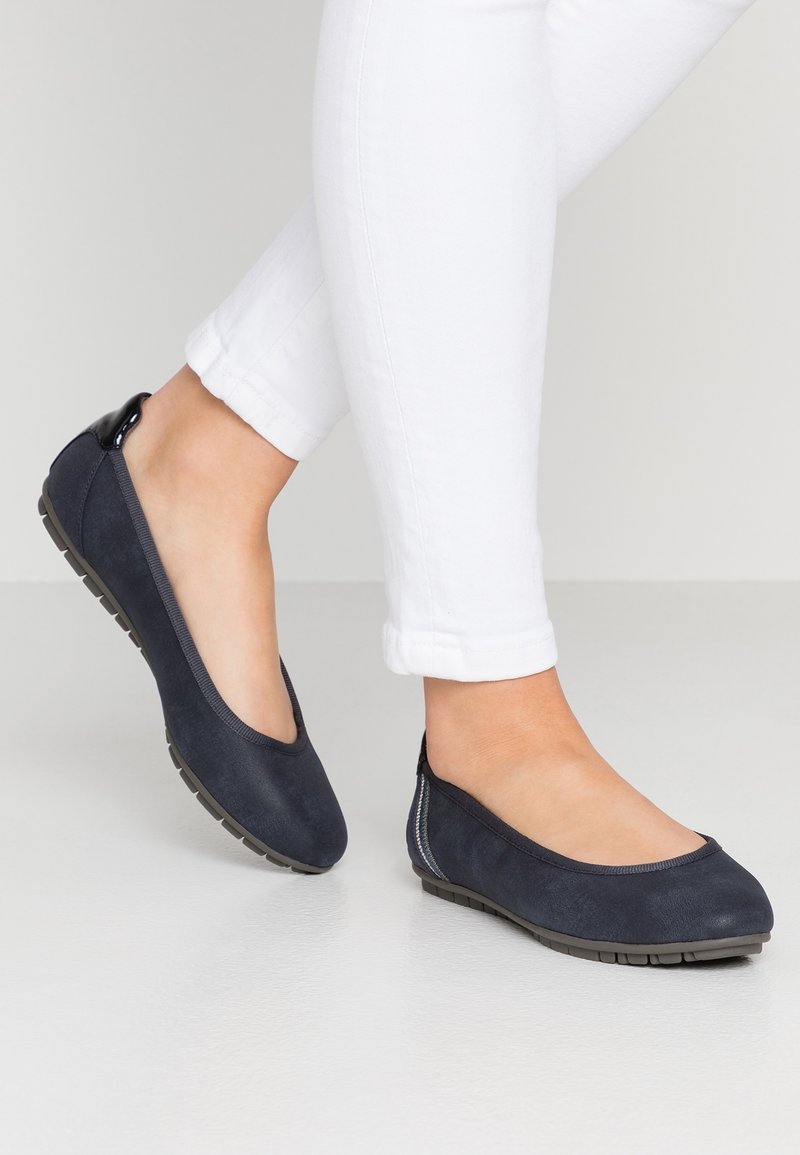 Pieds portant des ballerines bleu marine avec semelles en caoutchouc texturées et pantalons blancs retroussés, sur un fond clair uni.
