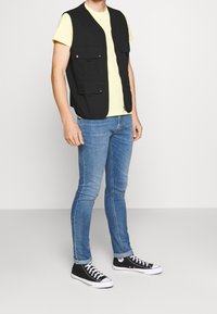 Svart ärmlös väst med fyra framfickor, bärs över en gul t-shirt, kombinerad med blå smala jeans och svarta lågskor.