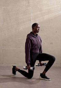 Two silver dumbbells held by a person in a dark purple cropped hoodie and black leggings, kneeling on one knee on a smooth floor.