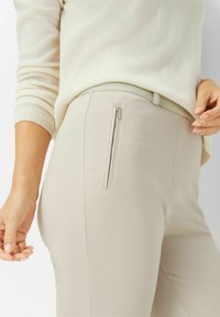 Beige, enganliegende Hose mit glatter Textur, die eine diskrete Reißverschlusstasche an der Seite und einen passenden Taillenbund aufweist.