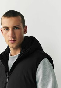 Schwarze gepolsterte Weste mit Kapuze, getragen über einem grauen Sweatshirt. Glatte Textur, Reißverschluss und verstellbare Kordeln an der Kapuze.