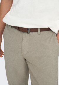 Pantalon gris à motifs avec un tissage texturé, doté d'une ceinture en cuir marron et d'un léger brillant. Présente des poches et une coupe ajustée.