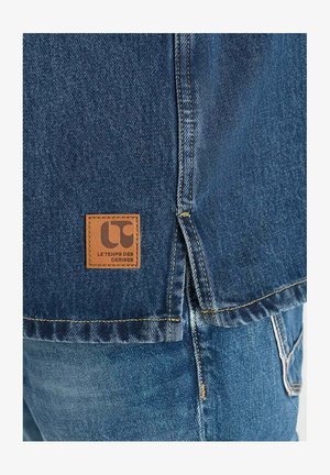 Denimjack met een geweven textuur, blauwe kleur en een leren merkpatch met de tekst "Le Temps des Cerises" op de achterkant.