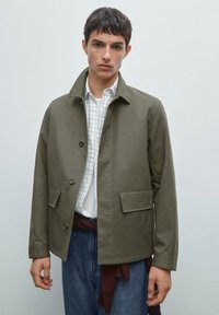Khaki jakke med knaplukning foran, klapkrave og to forlommer. Båret sammen med en hvid ternet skjorte og denim jeans.