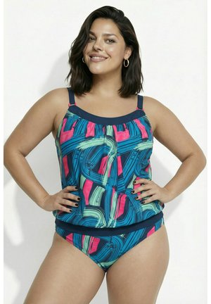Mujer sonriendo con las manos en las caderas, vistiendo un tankini de colores azul, rosa y verde con estampado, sobre un fondo liso.