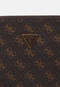 Brauner Geldbeutel aus synthetischem Leder mit einem wiederholten goldenen Logo-Muster, goldfarbener Dreieckslogo mit "GUESS USA" und gestickten Details.