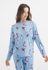 Dlouhý rukáv modrého pyžama se vzorem tučňáků lyžujících v růžové a žluté barvě, vyrobeno z měkké látky s uvolněným střihem.
